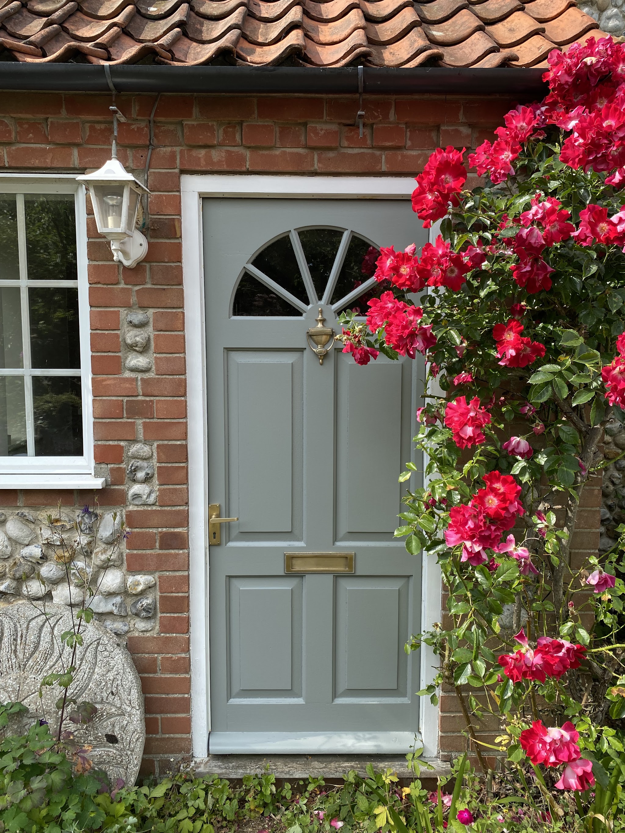 Sage grey panelled front door with a fanlight on a brick and flint cottage, red roses beside it