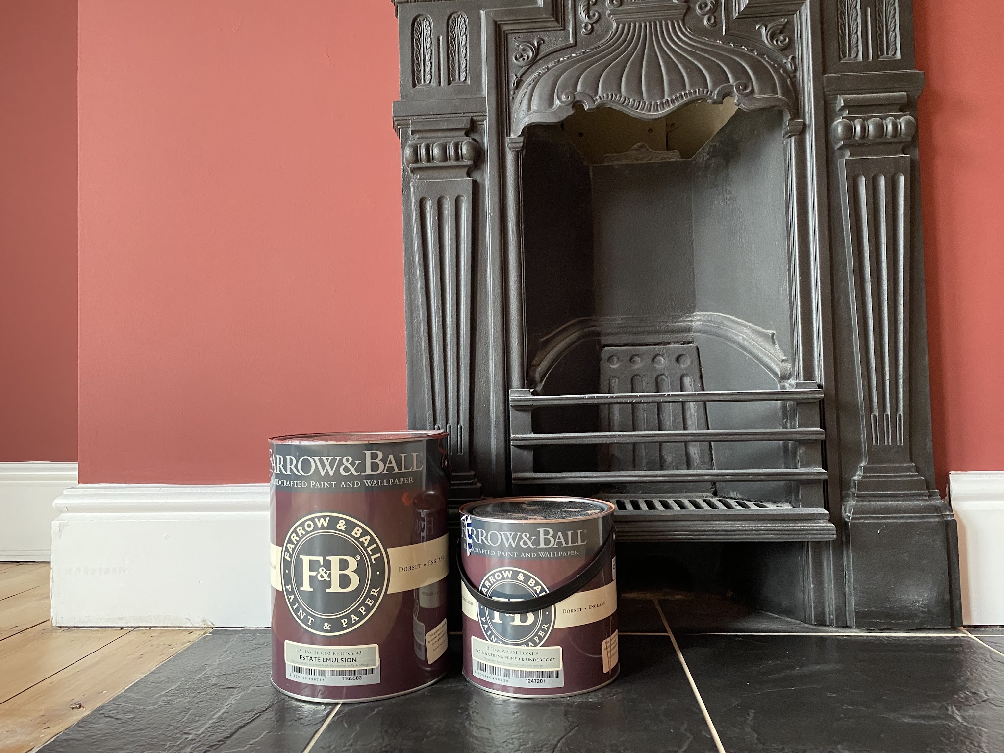 Terracotta painted wall and crisp white skirting around a cast iron fireplace, paint tins on the hearth